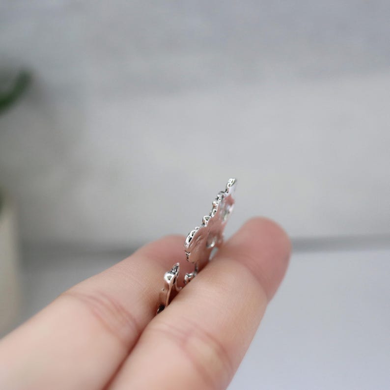 May include: A silver-colored, ornate metal charm with a detailed floral design. The charm is held between fingers, showcasing its intricate patterns and reflective surface. The charm's edges are slightly irregular, adding to its unique aesthetic.