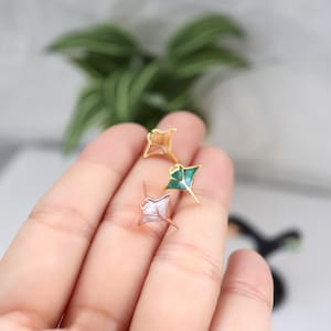May include: Three stud earrings shaped like manta rays, each featuring a metallic gold-tone outline. One earring has a light pink inlay, one has a vibrant green inlay, and the third has a pearlescent white inlay, all with a post back design.