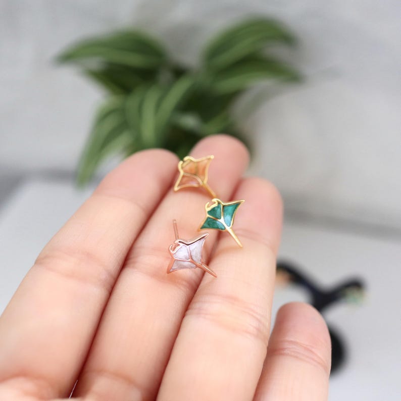 May include: Three stud earrings shaped like manta rays, each featuring a metallic gold-tone outline. One earring has a light pink inlay, one has a vibrant green inlay, and the third has a pearlescent white inlay, all with a post back design.