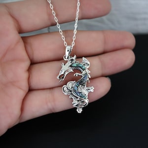 May include: A silver dragon pendant necklace with iridescent abalone shell inlay. The dragon is intricately detailed, with a curled body and open mouth. The necklace chain is delicate and silver. The pendant is approximately 2 inches in length.
