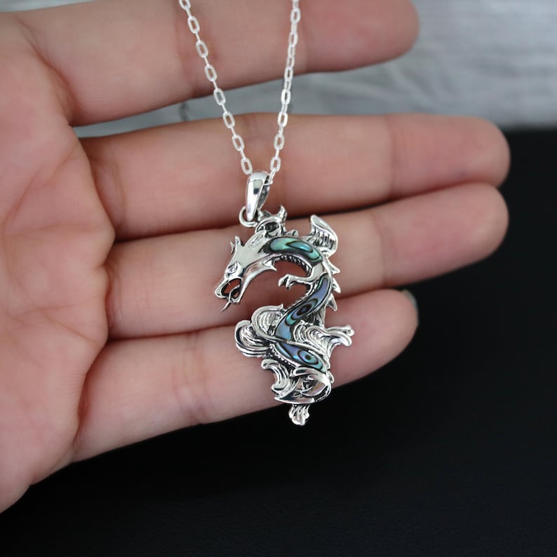 May include: A silver dragon pendant necklace with iridescent abalone shell inlay. The dragon is intricately detailed, with a curled body and open mouth. The necklace chain is delicate and silver. The pendant is approximately 2 inches in length.