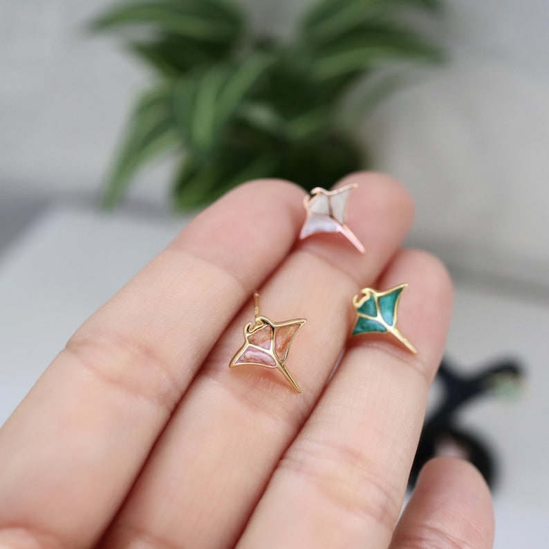 May include: Three small stingray-shaped stud earrings with gold-toned metal outlines. Each earring features a different colored inlay: one white, one peach-pink, and one teal-green. The stingray designs have a pointed tail and wide wings.