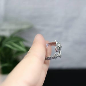 K&ouml;nnte beinhalten: Ein silberfarbener Ring mit einem einzigartigen Design. Der Ring hat ein strukturiertes, offenes Band mit einem detaillierten Pantherkopf und -schwanz. Der Kopf des Panthers hat ein gepunktetes Muster, der Schwanz ein strukturiertes Design.