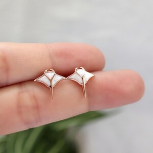 May include: A pair of stud earrings shaped like manta rays, featuring rose gold-toned metal outlines and white, textured inlay sections that form the wings and body of the sea creature, with a pointed tail extending from the center base.