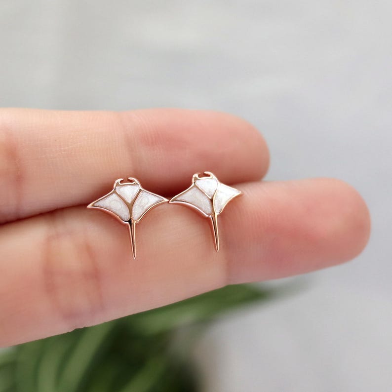 May include: A pair of stud earrings shaped like manta rays, featuring rose gold-toned metal outlines and white, textured inlay sections that form the wings and body of the sea creature, with a pointed tail extending from the center base.