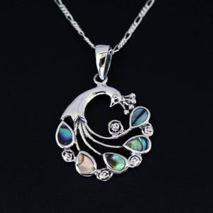 Puede incluir: Un collar con colgante de pavo real plateado con detalles de lágrima de abulón iridiscente. El colgante presenta un diseño detallado de pavo real con un cuerpo curvo y una cadena delicada. Las lágrimas de abulón son en tonos azules, verdes y rosas.