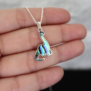 Könnte beinhalten: Eine silberne Halskette mit einem Dinosaurier-Anhänger. Der Anhänger hat die Form eines Brontosaurus und ist mit schillerndem Abalone-Muschel in Blau-, Grün- und Lilatönen eingelegt. Die Halskette ist filigran und silberfarben.
