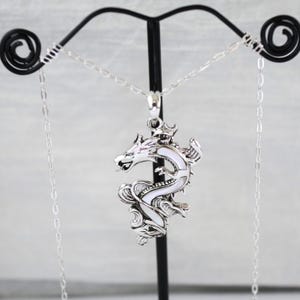 May include: A silver-colored dragon pendant necklace with a white enamel inlay. The dragon is detailed with scales and a crown, suspended from a delicate chain. The necklace is displayed on a black stand against a light background.