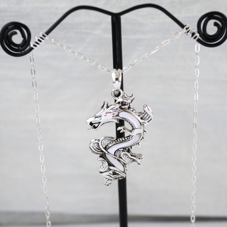 May include: A silver-colored dragon pendant necklace with a white enamel inlay. The dragon is detailed with scales and a crown, suspended from a delicate chain. The necklace is displayed on a black stand against a light background.