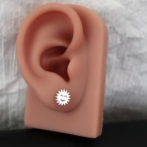 May include: A silver-colored stud earring in the shape of a smiling sun. The earring is displayed on a flesh-toned ear model against a white and gray background. The sun has a smiling face and spiky rays.