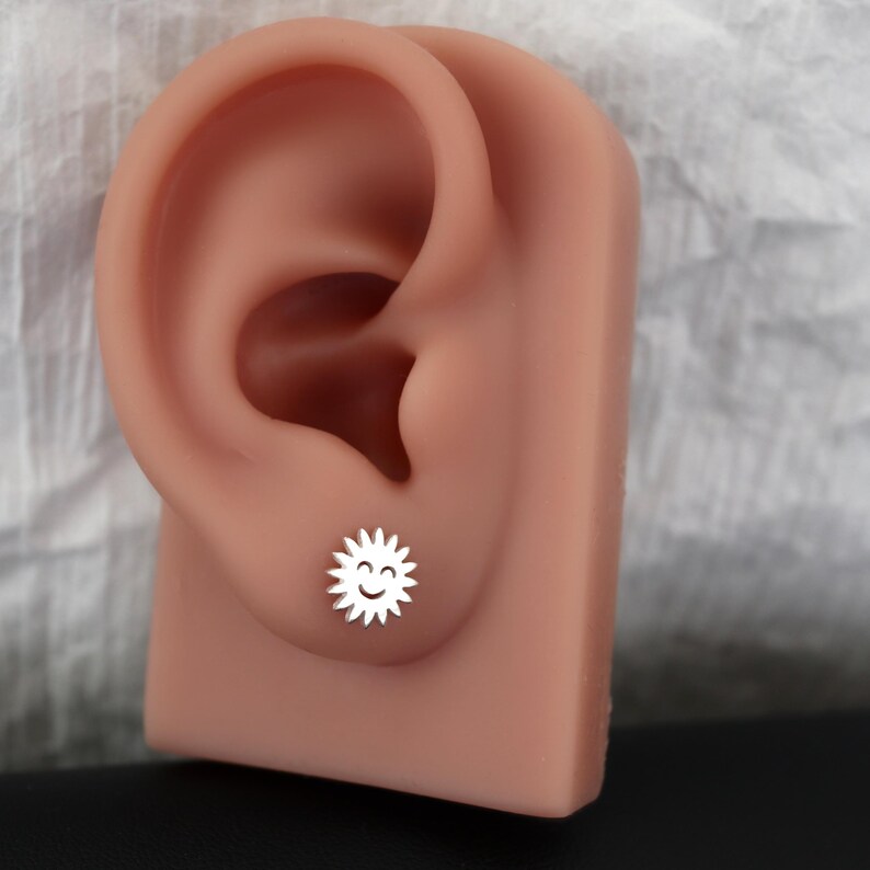 May include: A silver-colored stud earring in the shape of a smiling sun. The earring is displayed on a flesh-toned ear model against a white and gray background. The sun has a smiling face and spiky rays.