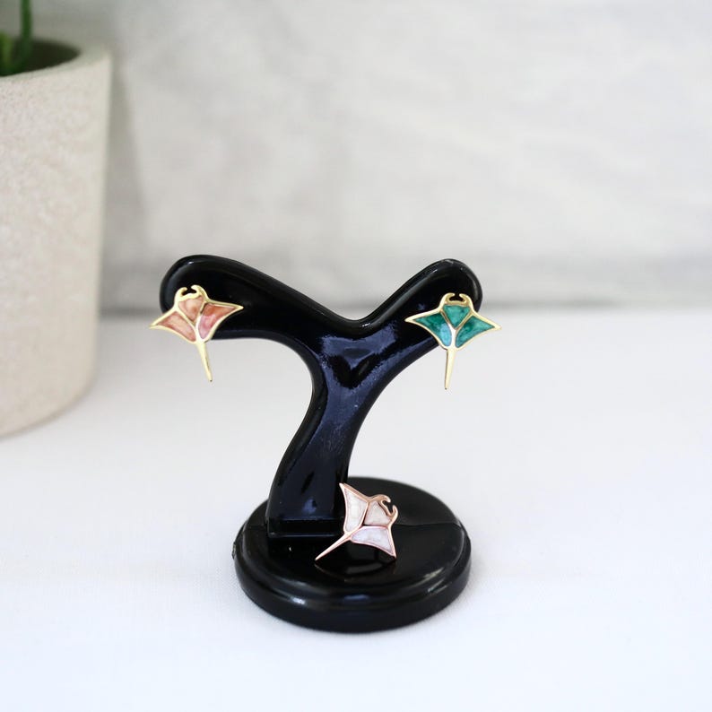 May include: Three stingray-shaped stud earrings displayed on a black stand. One earring features a pink marbled inlay, one has a vibrant teal marbled inlay, and the third is a white pearlescent inlay. Each piece has a gold-toned metal outline.