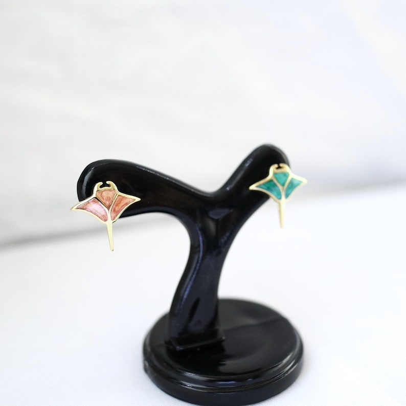 May include: A pair of stud earrings shaped like manta rays, featuring gold-toned metal outlines. One earring has a soft pink enamel inlay, while the other features a vibrant teal enamel inlay, both displayed on a black plastic stand.