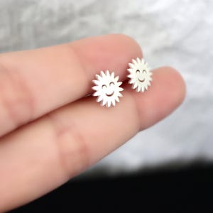 May include: A pair of small, silver-colored stud earrings. Each earring features a smiling sun design with spiky rays. The earrings are shown against a blurred background.