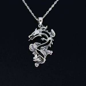 May include: A silver dragon pendant necklace. The dragon is intricately detailed with a curved body, open mouth, and a long, slender tongue. The pendant hangs from a delicate silver chain against a black background.