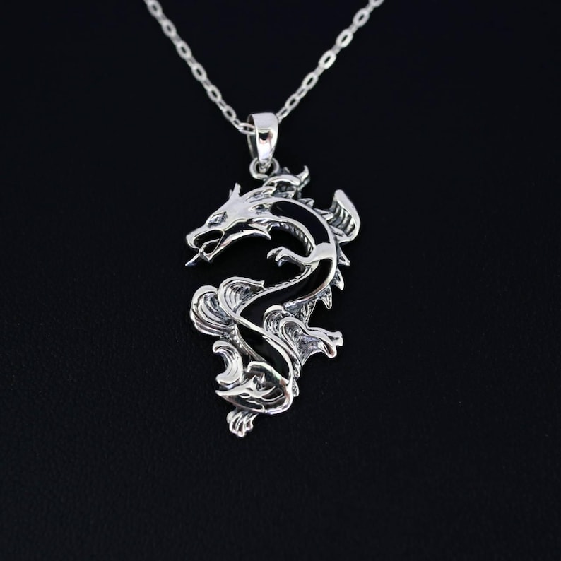 May include: A silver dragon pendant necklace. The dragon is intricately detailed with a curved body, open mouth, and a long, slender tongue. The pendant hangs from a delicate silver chain against a black background.