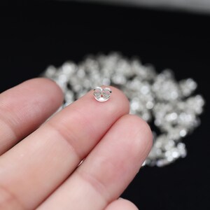 May include: A close-up of a silver earring back, also known as a butterfly clutch, held between fingers. The earring back is small, round, and has a butterfly-shaped design. A blurred background shows more earring backs.