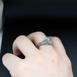 K&ouml;nnte beinhalten: Ein silberner Ring in Form eines Delfins mit strukturierter Oberfl&auml;che und kleinen schwarzen Akzenten. Der Ring wird an einem Finger getragen und zeigt sein detailliertes Design und das maritime Thema.
