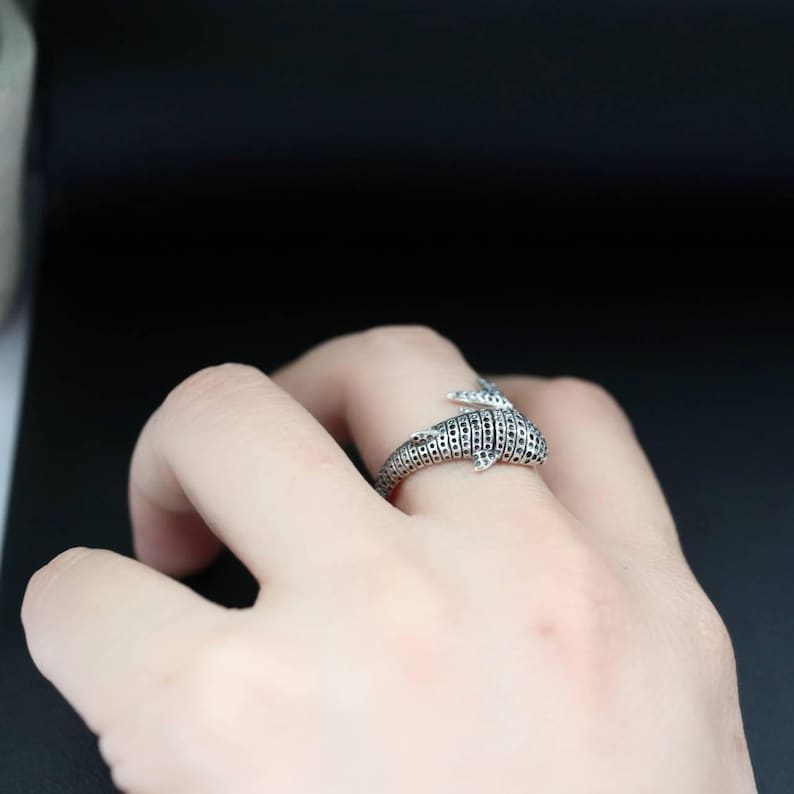 K&ouml;nnte beinhalten: Ein silberner Ring in Form eines Delfins mit strukturierter Oberfl&auml;che und kleinen schwarzen Akzenten. Der Ring wird an einem Finger getragen und zeigt sein detailliertes Design und das maritime Thema.