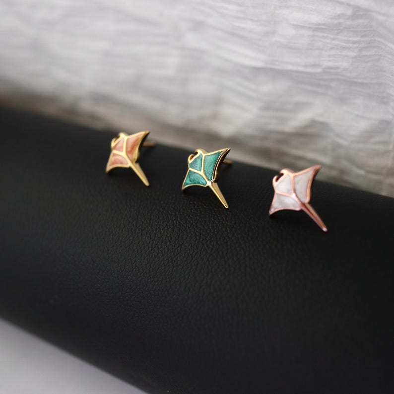 May include: Three stud earrings shaped like manta rays, each featuring a gold-toned metal outline. The left earring has a peach-colored inlay, the center earring has a teal-colored inlay, and the right earring has a white pearlescent inlay.