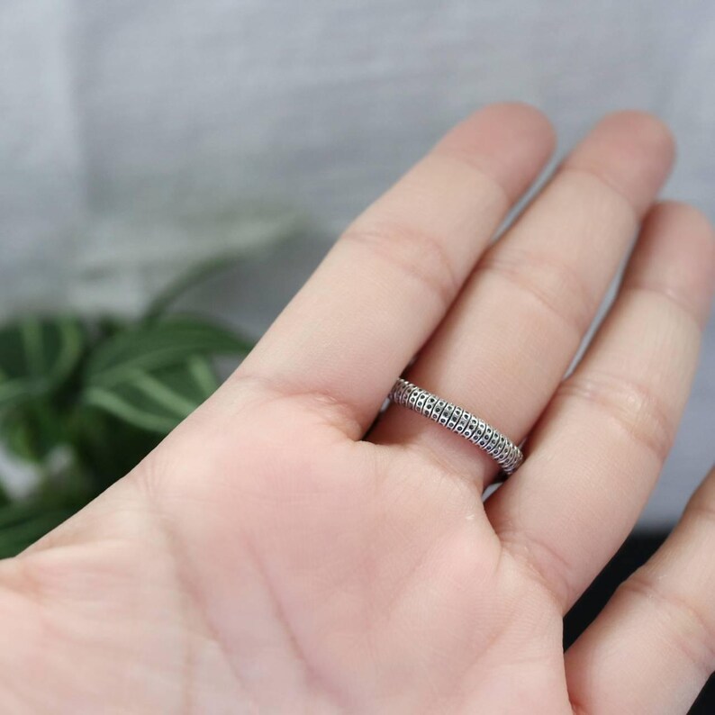 K&ouml;nnte beinhalten: Ein silberner Ring mit einem strukturierten, sich wiederholenden Muster. Der Ring wird auf einer Hand gezeigt, wobei der Fokus auf dem Design des Rings liegt. Das Ringband ist etwa 0,64 cm breit.