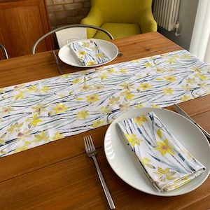 Puede incluir: Una mesa de madera con una silla amarilla y un camino de mesa blanco con un patrón floral. Dos platos blancos con servilletas florales a juego están colocados en la mesa. Un tenedor y un cuchillo están en la mesa.