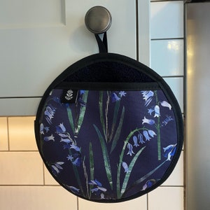 Puede incluir: Un agarrador de olla redondo azul y verde con un patrón floral de campanillas. El agarrador de olla tiene un bolsillo de tela negro y un lazo negro para colgar.