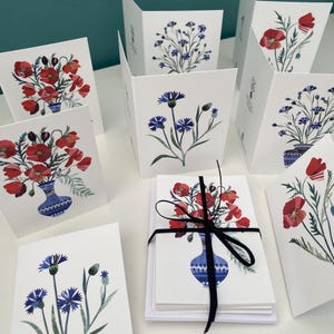 Puede incluir: Un juego de tarjetas de felicitación en blanco con ilustraciones de acuarela de amapolas rojas y aciano azul en jarrones azules y blancos. Las tarjetas están empaquetadas en una pila con una cinta negra.