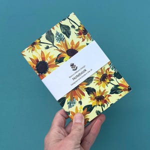 Puede incluir: Un cuaderno con un diseño de girasoles. La portada presenta girasoles amarillos con centros marrones y hojas verdes sobre un fondo amarillo claro. Una banda blanca envuelve el cuaderno con el texto "Bianco Williams Design Notebook Designed and Made in the UK."