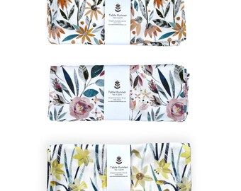 May include: Three rolled table runners with floral patterns. The top runner features orange and yellow flowers with green leaves. The middle runner has pink and blue flowers. The bottom runner displays yellow daffodils. Each runner is wrapped with a paper band.