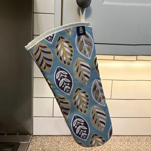 May include: A blue oven mitt with a white trim and a pattern of gold and brown leaves. The mitt is hanging from a hook on a white tiled wall.
