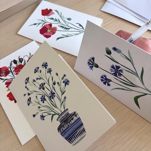 May include: Greeting cards with floral watercolor designs. One card features red poppies, while others showcase blue cornflowers in a vase and on their own. The cards are on a light beige background.