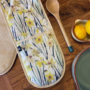Puede incluir: Un guante de cocina doble con un estampado floral de narcisos en amarillo, blanco y verde. El guante tiene un borde y un bolsillo color crema. También se ve una cuchara de madera con mango turquesa y un cuenco con limones.