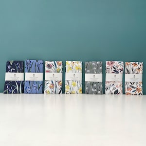 Könnte beinhalten: Acht Geschirrtücher mit Blumendesigns. Die Handtücher sind in einer Reihe auf einer weißen Oberfläche angeordnet. Jedes Handtuch hat ein anderes Blumenmuster, darunter Blausternchen, Narzissen und Schneeglöckchen. Die Handtücher sind alle weiß mit einem schwarzen Rand und dem Aufdruck "Luxury Tea Towel".