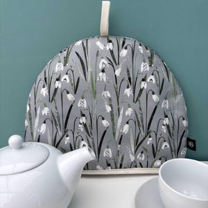 Può includere: Una teiera grigia con un motivo floreale di fiori di bucaneve bianchi e steli verdi. La teiera ha un bordo color crema e un anello color crema. Una teiera e una tazza bianche sono in primo piano.