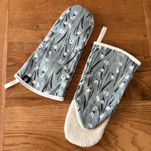 Op de afbeelding: Een paar ovenwantjes met een grijze achtergrond en een wit bloemenpatroon. Een wantje heeft een terrycloth voering, terwijl de andere een witte rand heeft.