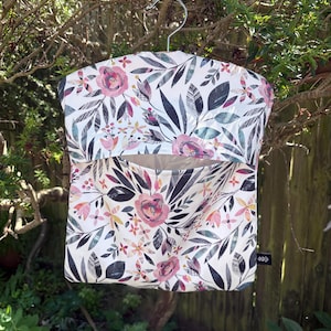 May include: A hanging fabric storage bag featuring a white background with a botanical print of pink roses, dark gray leaves, and small yellow flowers. The bag has a wide horizontal opening across the center and is suspended by a metal hanger.