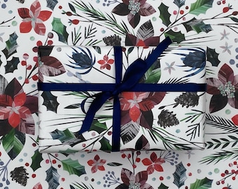 May include: A white gift wrap with a festive design featuring red and green poinsettia flowers, holly leaves, and pine needles. A navy blue ribbon is tied around the gift wrap.