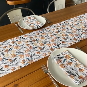 Op de afbeelding: Een witte tafelrunner met een bloemenpatroon in tinten oranje, blauw en grijs. De runner ligt op een houten tafel met twee couverts, elk met een wit bord en een bijpassende bloemen servet.