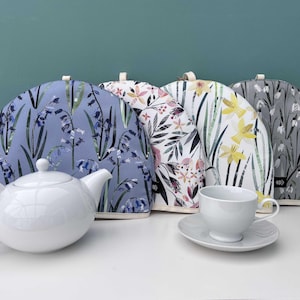 Può includere: Una teiera, una tazza e un piattino in ceramica bianca sono esposti con quattro copriteiere. I copriteiere presentano motivi floreali blu, rosa, gialli e grigi. Lo sfondo è di colore verde acqua.
