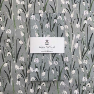 Könnte beinhalten: Ein graues Geschirrtuch mit einem weißen Schneeglöckchen-Blütenmuster. Das Handtuch hat ein Etikett mit der Aufschrift "Luxury Tea Towel, Designed and Made in the UK, 100% Cotton, www.BiancaWilliamsDesign.com".