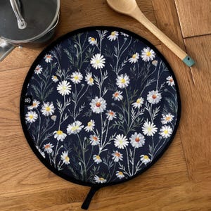 May include: Circular navy blue pot stand with a black edge and a hanging loop. It displays a floral design of white daisies with yellow centres and green stems. A wooden spoon is on the wooden surface.