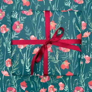 Op de afbeelding: Een rechthoekige geschenkdoos verpakt in donkergroenblauw papier met een herhalend patroon van rode klaprozen en groen gebladerte. De doos is gestrikt met een glad, donkerrood satijnen lint dat in het midden tot een strik is geknoopt.