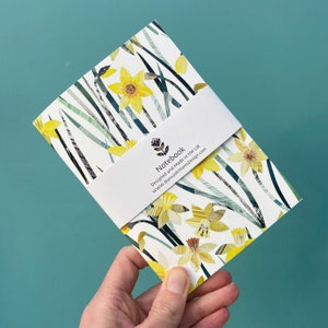 May include: A white notebook with a yellow daffodil floral pattern. The cover has a white label with the text "Notebook" and "Designed and Made in the UK" and "www.BiancaWilliamsDesign.com".
