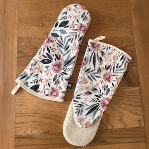 May include: A pair of oven gloves with a floral pattern. The gloves are white with pink and blue flowers and leaves. One glove has a terry cloth lining.
