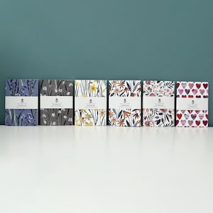 Puede incluir: Seis cuadernos con diferentes patrones y una etiqueta blanca que dice "Notebook" en cada uno. Los cuadernos presentan diseños florales en tonos azules, grises, amarillos, naranjas, rosas y rojos.