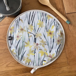 May include: Circular pot stand featuring a daffodil pattern on a white background. It has a cream-coloured border and a hanging loop. A wooden spoon and a pot are visible in the background.