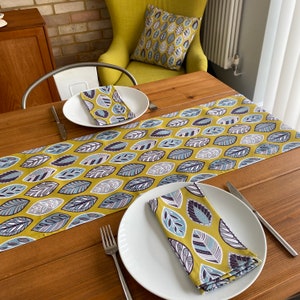 May include: A yellow table runner with a blue and grey leaf pattern. The runner is on a wooden table with two white plates and napkins with the same leaf pattern.