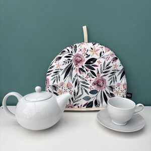 Puede incluir: Un juego de té de cerámica blanca con tetera, taza y platillo, y un calentador de té floral. El calentador de té presenta un fondo blanco con patrones florales rosas y morados. El juego está dispuesto sobre una superficie blanca.