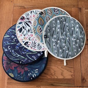 Op de afbeelding: Set van vijf ronde pottenlapjes met verschillende bloemenpatronen. De pottenlapjes zijn gemaakt van stof met een witte rand en hebben een lus om op te hangen. De patronen omvatten blauwe bellen, sneeuwklokjes en andere bloemenpatronen.